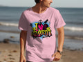 On the beach, someone wears a light pink T-shirt by Garment Graphics, featuring a vibrant I ♥️ Graffiti graffiti design. This street art-inspired piece embodies the spirit of Graffiti Love T-Shirts, with waves visible in the background.