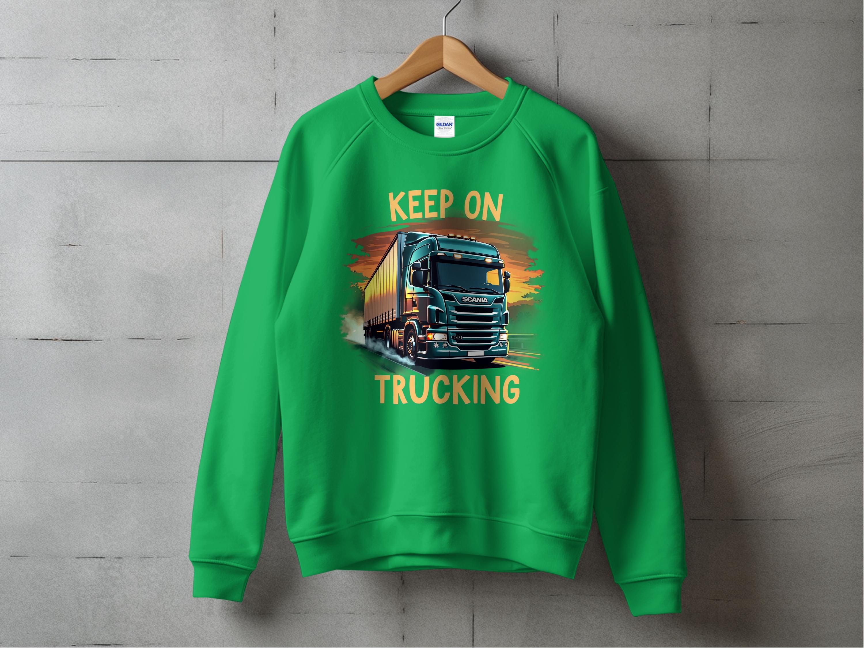 A bold truck-themed graphic sweatshirt by Garment Graphics hangs on a textured wall, showcasing a large truck image with Keep On Trucking printed above and below, embodying an adventurous spirit.