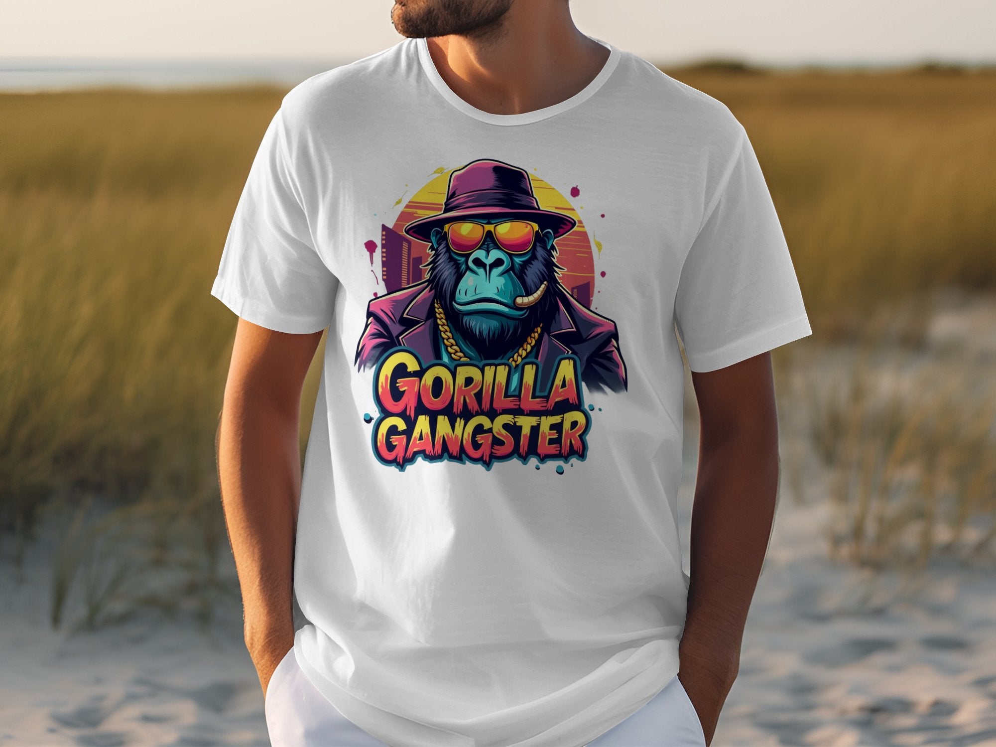 A person sports a striking Graphic T-Shirt from Garment Graphics, featuring a gorilla dressed as a gangster with sunglasses, hat, and cigar. The words Gorilla Gangster appear in colorful letters beneath the image against a grassy backdrop.