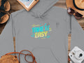 From Garment Graphics, a gray hoodie with Take It Easy in neon blue and yellow is on wood, surrounded by a hat, sunglasses, notebook, sandals, map, camera, and woven bag—ideal for comfy style.