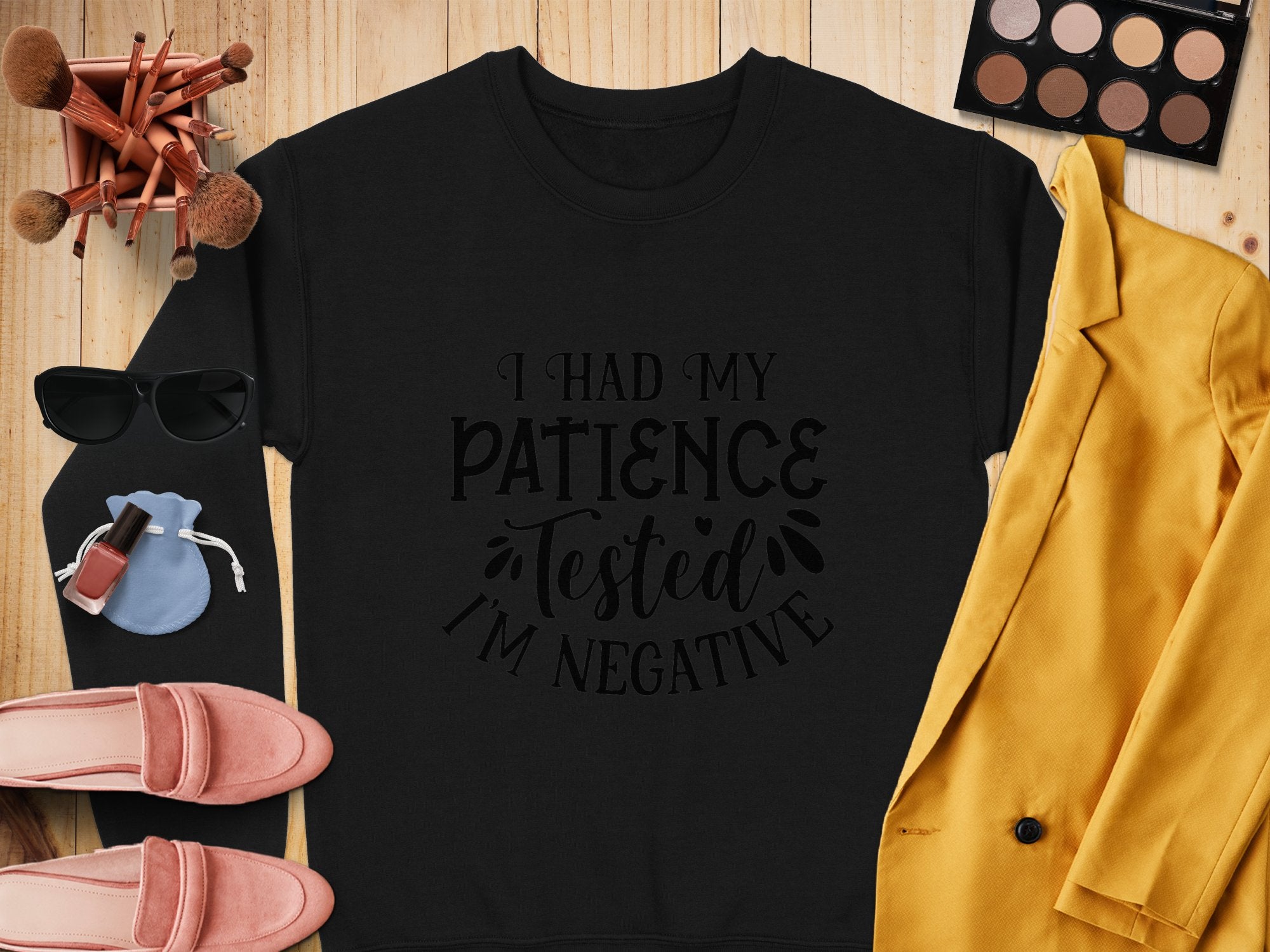 A flat lay showcasing a Garment Graphics sweatshirt with the phrase I had my patience tested Im negative, paired with a mustard yellow blazer, pink loafers, sunglasses, makeup brushes, a light blue pouch, nail polish, and an eyeshadow palette on wood—a fun choice for casual outings.