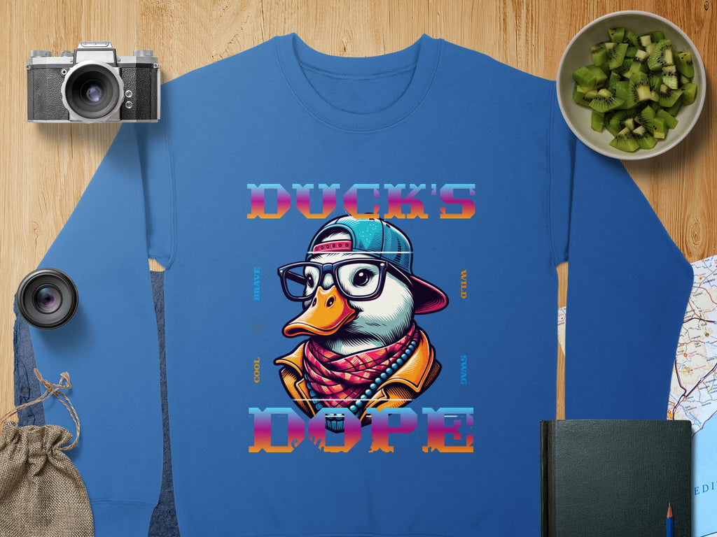 Blue sweatshirt by Garment Graphics, displaying a trendy cartoon duck wearing glasses, a hat, and a scarf. The design is complemented by the phrase Ducks Dope in vibrant letters. Nearby are artfully arranged items: camera, lens, sliced kiwi in bowl, bag, book, and map.