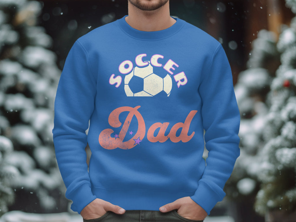 A person is wearing a stylish blue Garment Graphics Soccer Dad sweatshirt featuring a soccer ball graphic above Dad. The unisex size ensures comfort for everyone, while blurred snow-covered trees in the background create a wintry atmosphere.