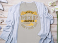 Displayed on a wooden surface, Garment Graphics gray t-shirt features the humorous quote, If hes got golf clubs in his truck or a camper in his driveway, I dont hire him, surrounded by a light cardigan, flowers, and accessories—an ideal gift for humor enthusiasts.