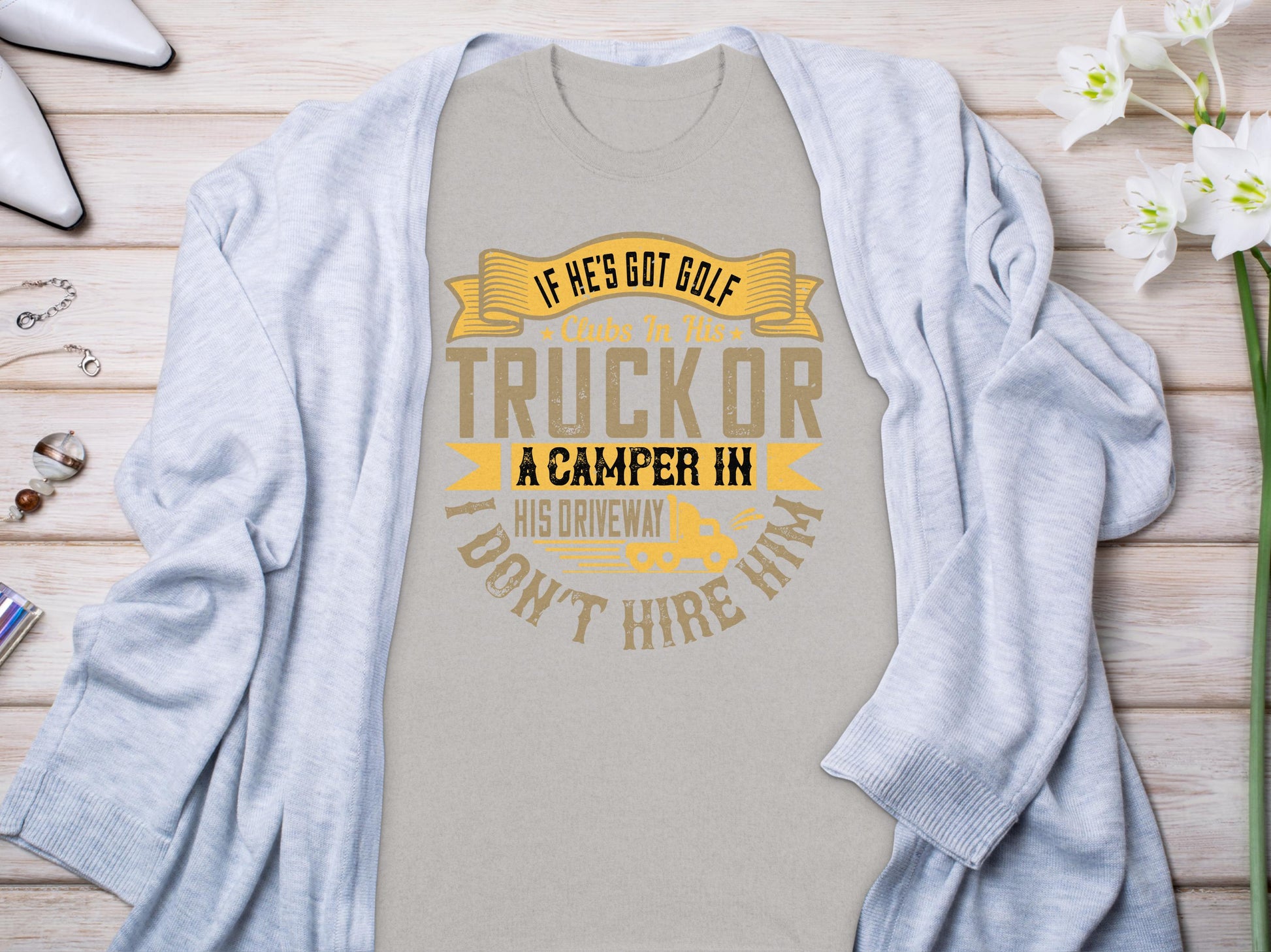 Displayed on a wooden surface, Garment Graphics gray t-shirt features the humorous quote, If hes got golf clubs in his truck or a camper in his driveway, I dont hire him, surrounded by a light cardigan, flowers, and accessories—an ideal gift for humor enthusiasts.