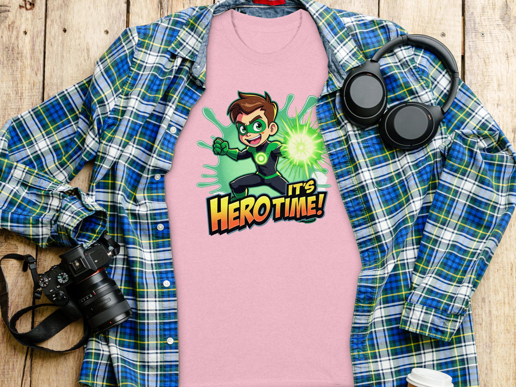 A vibrant light pink t-shirt from Garment Graphics, showcasing a superhero cartoon with green gloves and a glowing aura, captioned Hero Time! is displayed on a blue-green plaid shirt beside a camera, headphones, and cup atop a wooden surface.