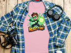 A vibrant light pink t-shirt from Garment Graphics, showcasing a superhero cartoon with green gloves and a glowing aura, captioned Hero Time! is displayed on a blue-green plaid shirt beside a camera, headphones, and cup atop a wooden surface.