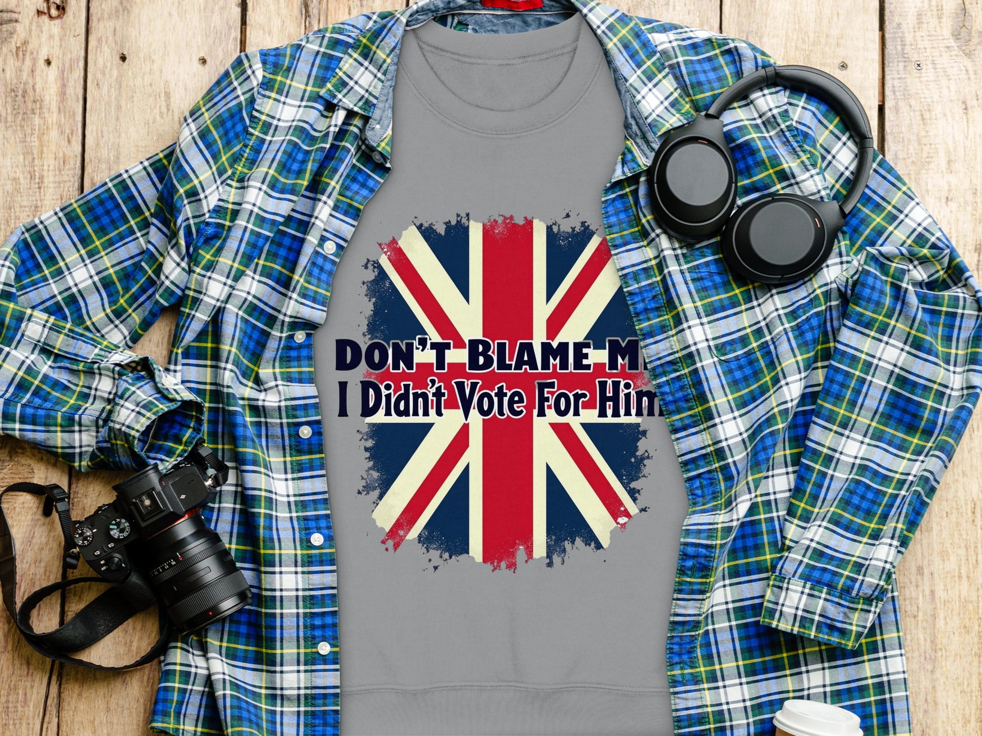 A gray unisex t-shirt from Garment Graphics features a British flag and the bold message Dont Blame Me, I Didnt Vote For Him. It is displayed on wood alongside a blue plaid shirt, camera, and headphones.