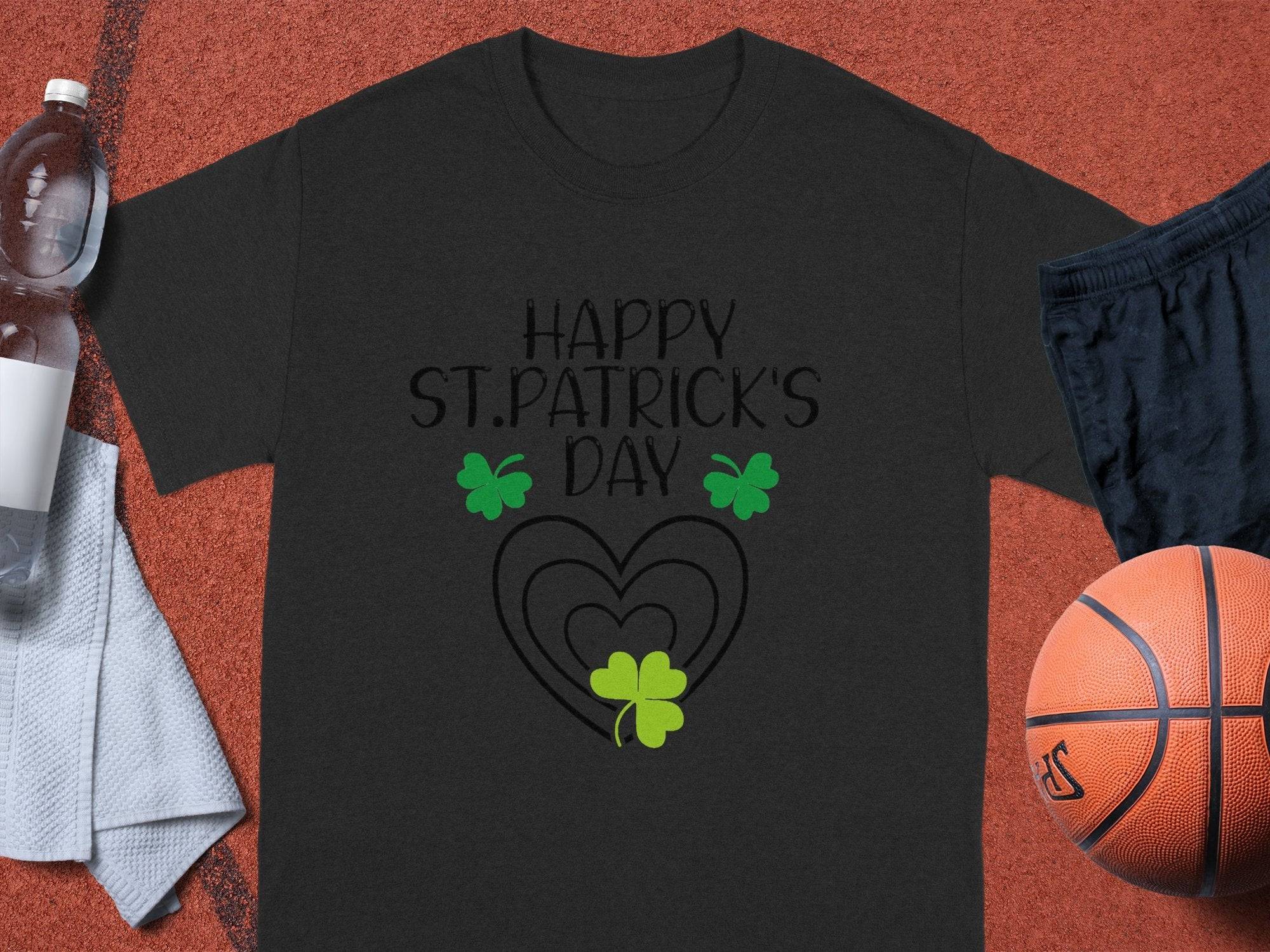A black Garment Graphics t-shirt with Happy St. Patricks Day and green shamrocks brings festive flair to your wardrobe. Set on an orange surface, it pairs with a basketball, water bottle, towel, and dark shorts for a lively look.