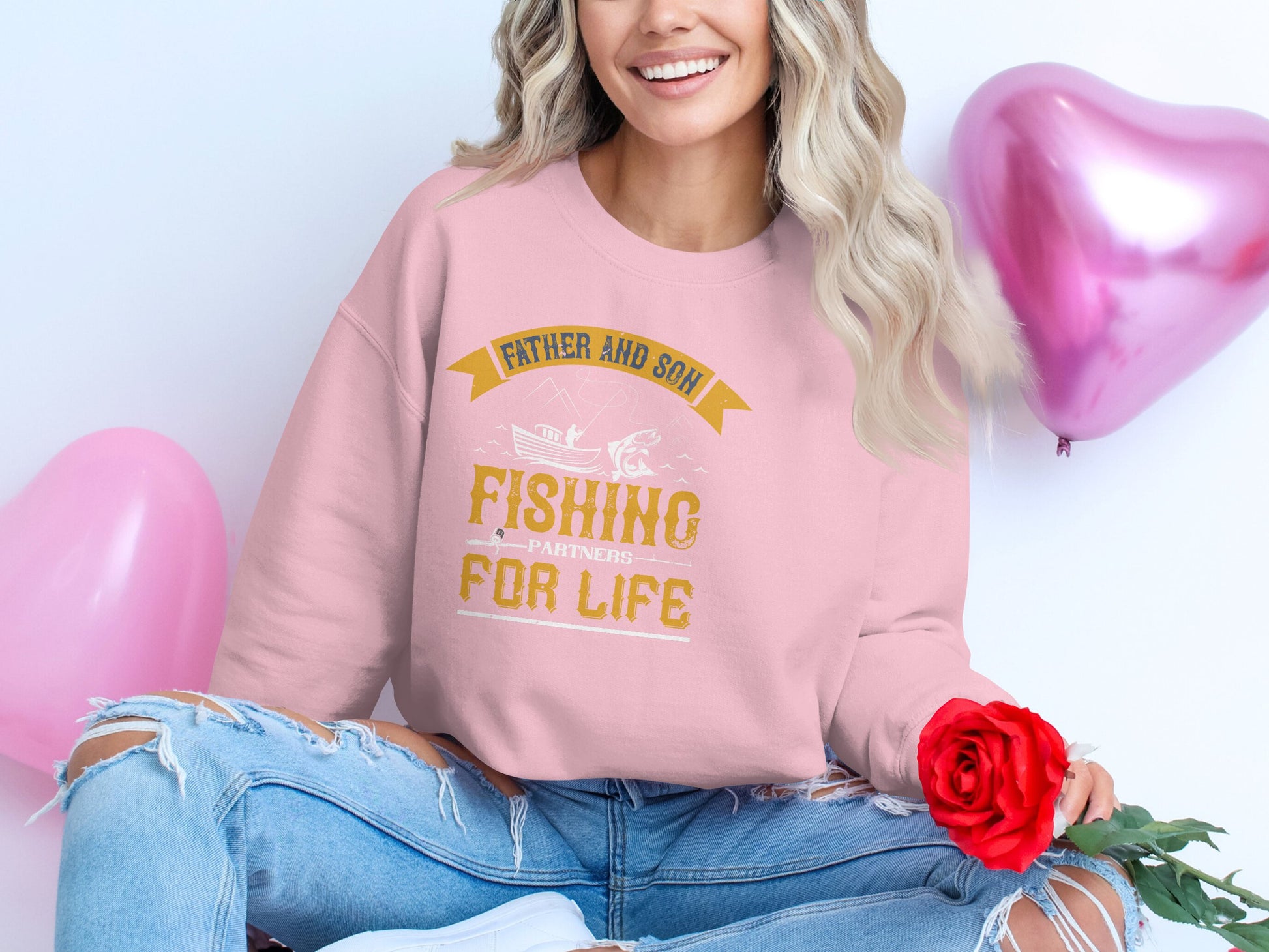 A woman smiles on the floor in a pink Garment Graphics sweatshirt that says “Father and Son Fishing Partners for Life.” She holds a red rose with heart-shaped balloons around her, and her ripped jeans add a casual touch to the heartfelt moment.