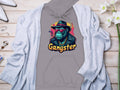 Garment Graphics offers a gray hoodie featuring a green monkey in gangster style with a black hat, sunglasses, gold chains, and a cigar. Gangster is printed in yellow below the image. Made from medium-heavy fabric, its displayed layered with a light cardigan on wood.