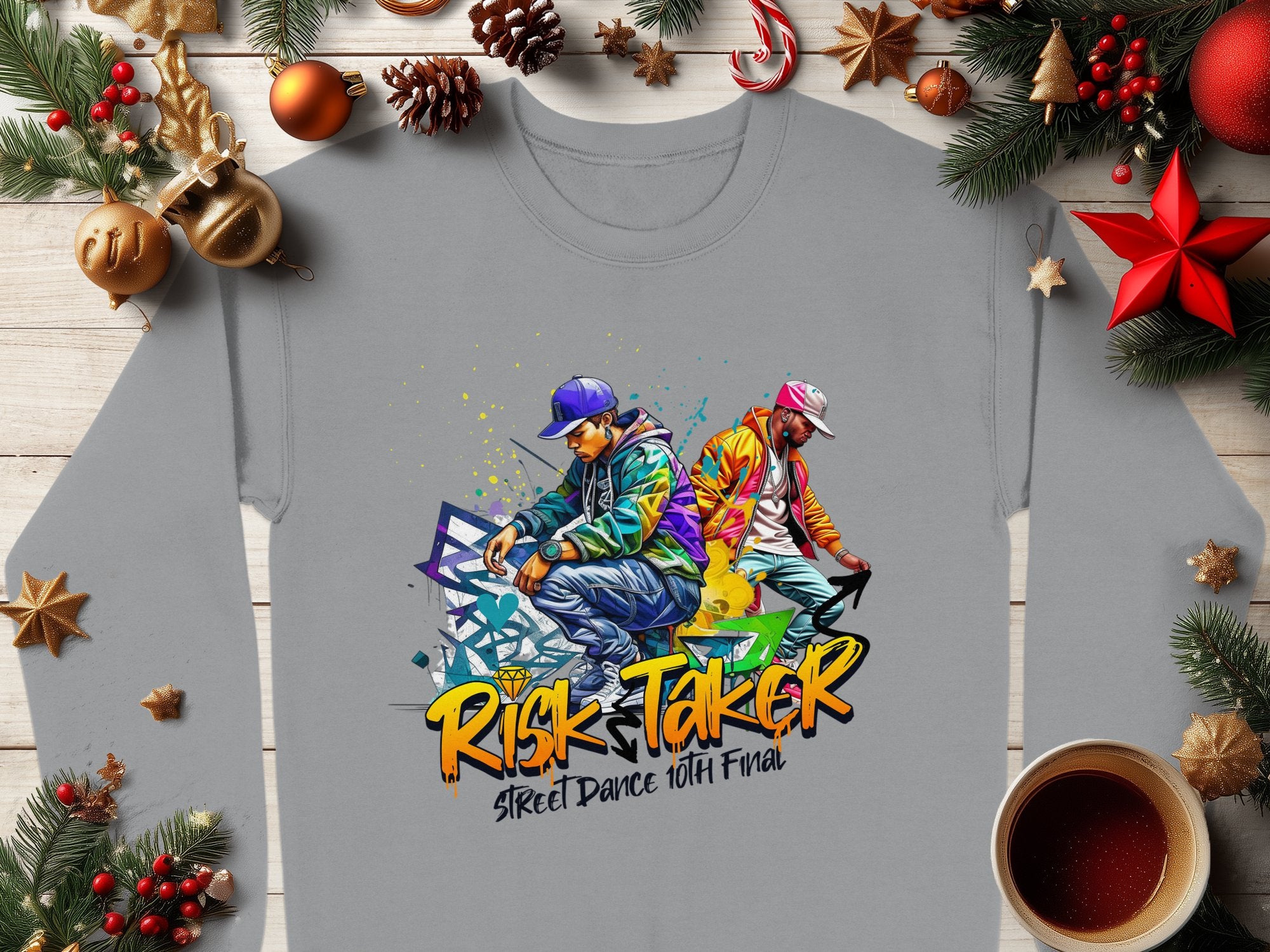Urban-style gray sweatshirt by Garment Graphics with street dance graphics of two dancers in colorful clothing, spray paint effects, and the text Risk Taker, Street Dance 10th Final. Unisex sizing. Displayed on a wooden surface with festive decor and a coffee cup.