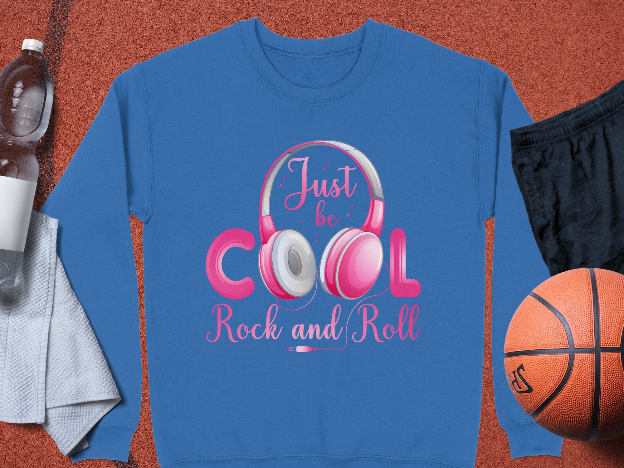 This stylish Garment Graphics blue sweatshirt features pink and white headphones with the phrase Just be COOL Rock and Roll in various fonts. Paired with black shorts, a basketball, water bottle, and towel, its ideal for music lovers on the go.