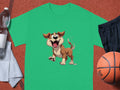 The Garment Graphics green T-shirt is ideal for dog lovers, showcasing a cartoon dog on a sports court. Nearby are a basketball, black shorts, a white towel, and a water bottle.