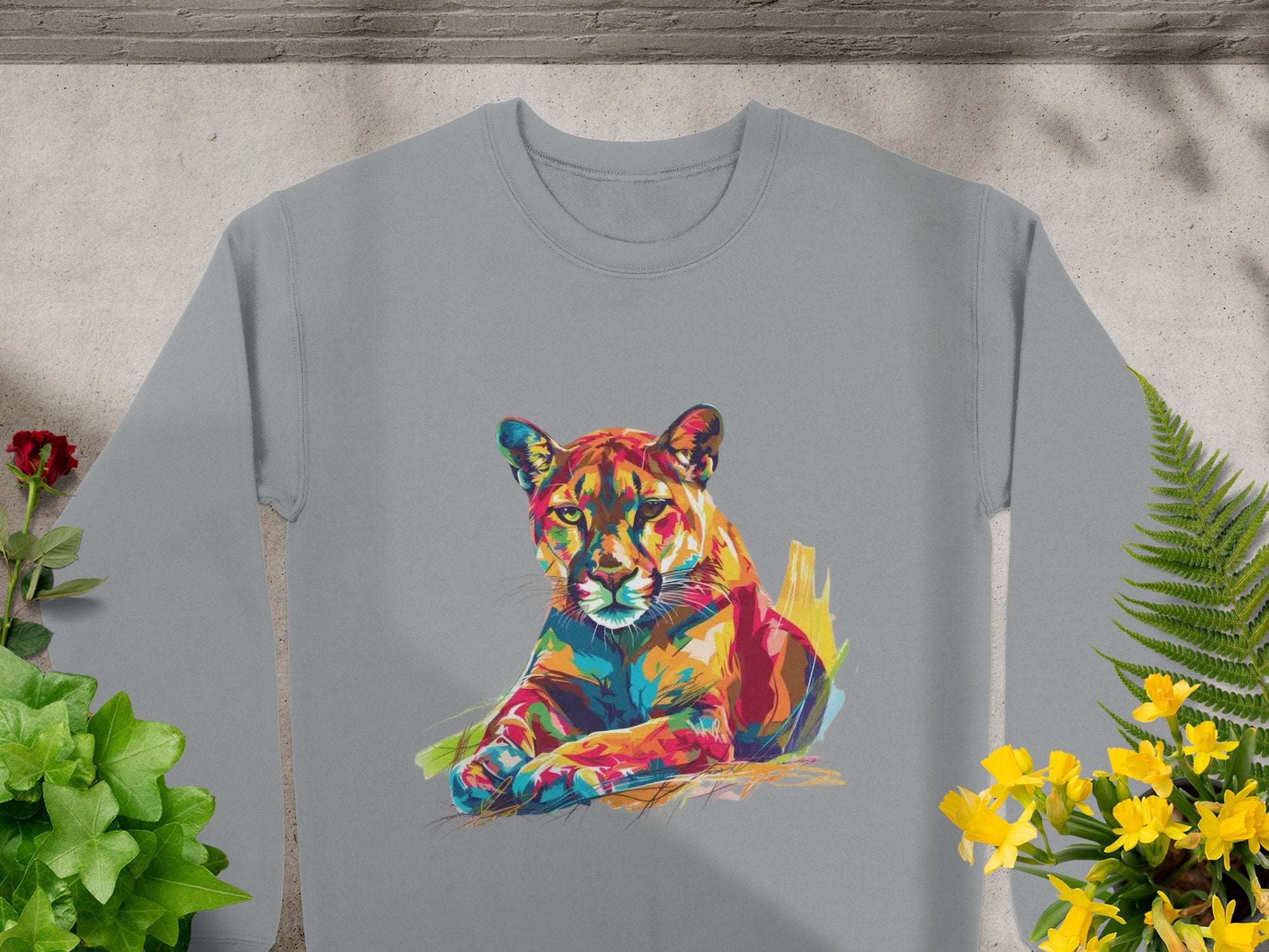 A Garment Graphics gray unisex sweatshirt with a vibrant artistic lion face design is laid flat on a light surface, surrounded by green plants and yellow flowers, highlighting its unique style.