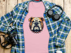 A pink Garment Graphics T-shirt featuring a bulldog design rests on a wooden surface. Its surrounded by stylish shirts, including a blue and green plaid one. Nearby essentials include a camera, headphones, a lidded white cup, and a smartphone.