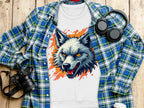 A white Garment Graphics T-shirt with a fierce wolf design is laid on a wooden surface, paired with a blue and green plaid shirt for unique flair. Nearby are black headphones, a camera, and a white coffee cup with a lid.