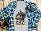 A Garment Graphics gray sweatshirt featuring an ornate eye design and GAZE is stylishly layered over a blue and green plaid shirt on wood. A camera, headphones, and white travel cup complete this unisex look.