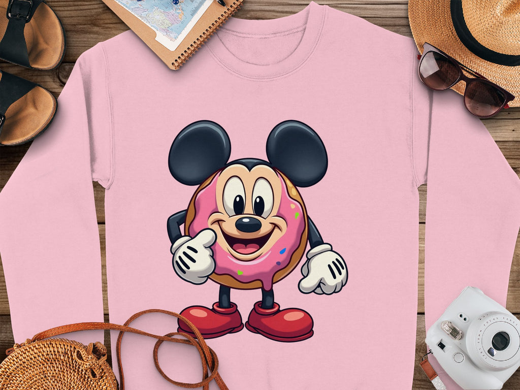 The Garment Graphics pink unisex sweatshirt features a whimsical donut character akin to an iconic mouse, laid on wood and styled with sandals, a journal, straw hat, sunglasses, and camera for playful adventures.