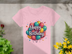 A vibrant 100% pink cotton T-shirt by Garment Graphics lies on a gray surface, featuring a colorful Happy Birthday design with balloons and stars. It is surrounded by lush green and yellow plants, with a small red flower enhancing the festive scene on the left.