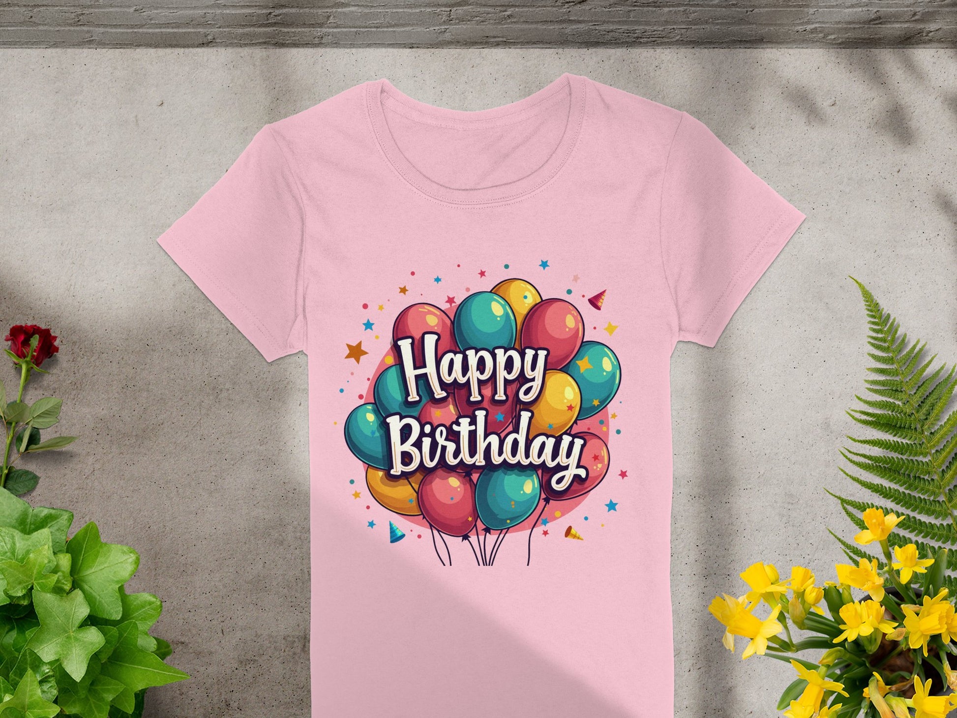 A vibrant 100% pink cotton T-shirt by Garment Graphics lies on a gray surface, featuring a colorful Happy Birthday design with balloons and stars. It is surrounded by lush green and yellow plants, with a small red flower enhancing the festive scene on the left.