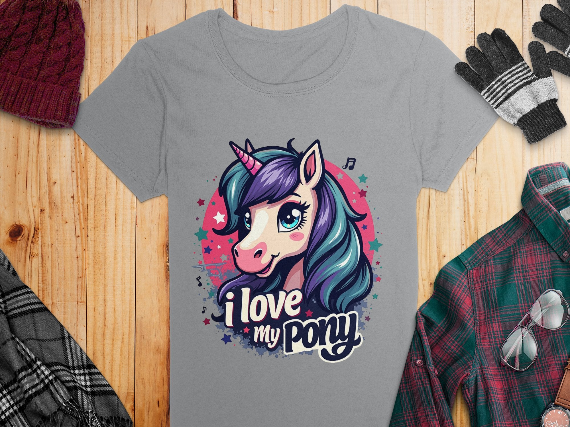 Garment Graphics gray t-shirt features a vibrant unicorn with a purple and blue mane, and I Love My Pony text. Ideal for unicorn lovers, its displayed on a wooden surface surrounded by winter clothes like gloves and a hat.