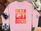 The Garment Graphics pink unisex sweatshirt reads Born to be Musician in vibrant orange and red, with drummer and guitarist silhouettes. Perfect for music lovers, its pictured with travel essentials like a camera, gloves, and binoculars—ideal for any adventure.