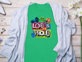 A Garment Graphics green t-shirt with Loud & Proud and phrases like Hey ho, lets go is displayed on wood. Ideal for football enthusiasts, its paired with a light gray cardigan and decorative items.