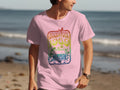 A music enthusiast in a pink Garment Graphics T-shirt, showcasing a vibrant Woodstock 1969 design with a retro van and psychedelic elements, walks along the sandy beach as waves roll in under the clear sky.