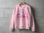 A Garment Graphics pink sweatshirt hangs on a wooden hanger against a concrete wall. It features Just be Cool Rock and Roll in vibrant pink and purple text, paired with stylish Rock and Roll Pink Headphones illustrations, making it a chic statement for music lovers.