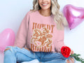 A smiling individual in a Garment Graphics Howdy Rodeo sweatshirt, ideal for rodeo fans, sits cross-legged. The pink top displays a cowgirl on horseback. Holding a red rose, they are surrounded by pink heart-shaped balloons, radiating cowboy charm.