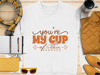 Flat lay image features Garment Graphics white shirt with Youre My Cup of Tea in brown text. Cozy accents include a plaid scarf, black coffee, unisex sizing, a notebook with pen, quilted jacket, yellow sunglasses, and clutch on a wooden surface.