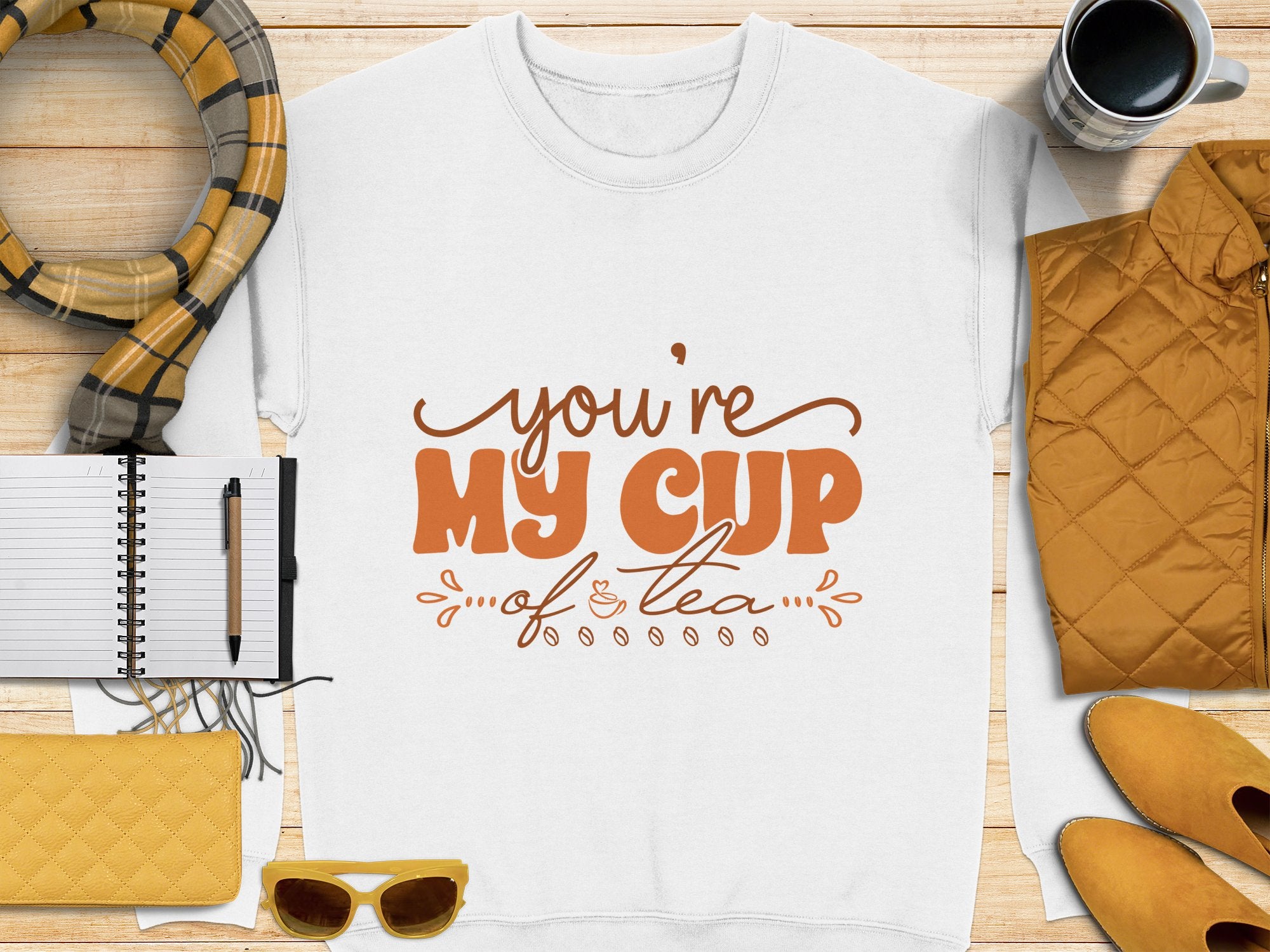 Flat lay image features Garment Graphics white shirt with Youre My Cup of Tea in brown text. Cozy accents include a plaid scarf, black coffee, unisex sizing, a notebook with pen, quilted jacket, yellow sunglasses, and clutch on a wooden surface.