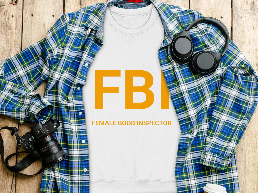 A flat lay image from Garment Graphics displays a white sweatshirt with FBI Female Boob Inspector in bold orange letters, accompanied by a blue plaid shirt, headphones, camera, and to-go coffee cup on wood. This playful piece offers unisex sizing for versatile style options.