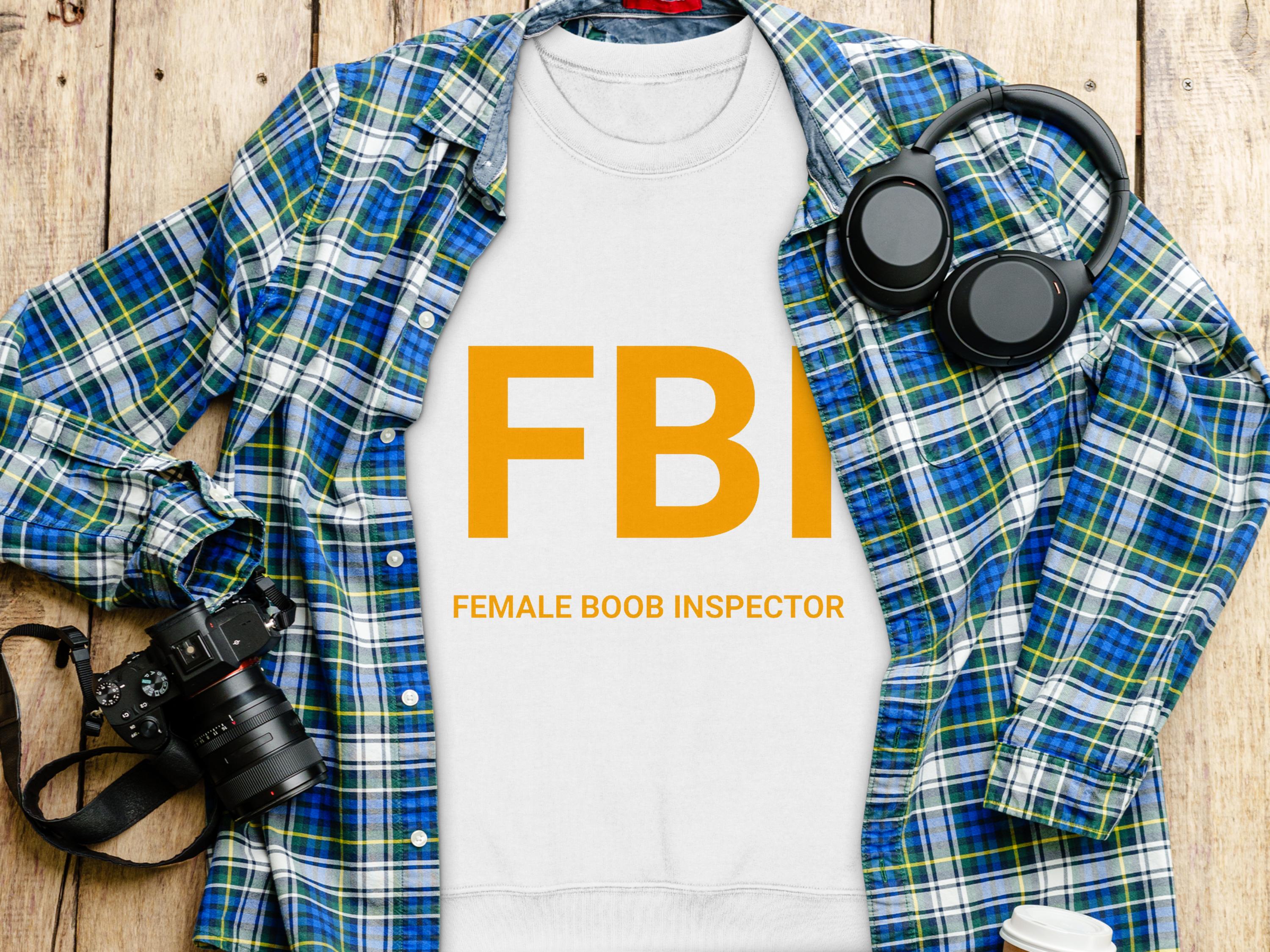 A flat lay image from Garment Graphics displays a white sweatshirt with FBI Female Boob Inspector in bold orange letters, accompanied by a blue plaid shirt, headphones, camera, and to-go coffee cup on wood. This playful piece offers unisex sizing for versatile style options.
