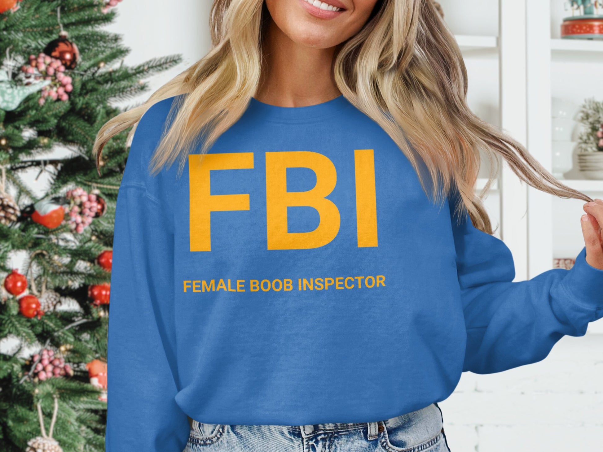 An individual wears a Garment Graphics graphic print sweatshirt reading FBI in bold yellow letters with FEMALE BOOB INSPECTOR beneath, standing indoors with unisex sizing near a decorated Christmas tree.