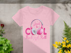 A pink Garment Graphics T-shirt, featuring a bold Just be cool, rock and roll design, is artfully displayed on a stone surface amongst lush greens and vibrant yellow flowers—perfect for stylish music lovers.