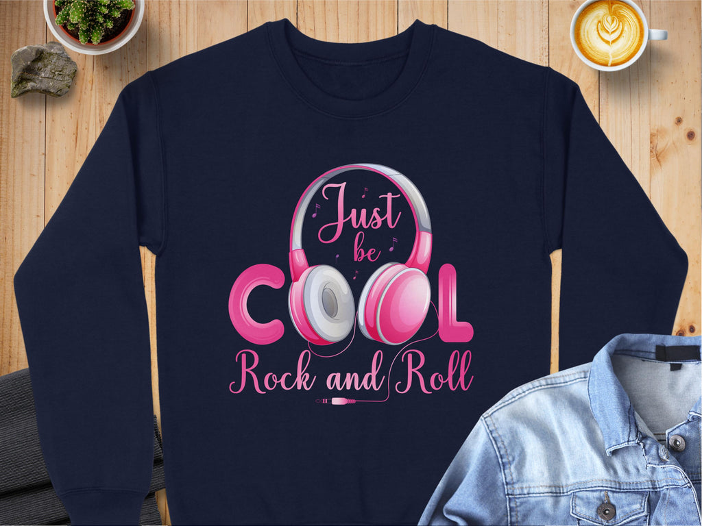 A navy sweatshirt by Garment Graphics features Rock and Roll Headphones and Just be COOL Rock and Roll text in pink and white. Elegantly placed on a wooden surface with a denim jacket, it’s perfect for fashion lovers, with a latte completing the look.