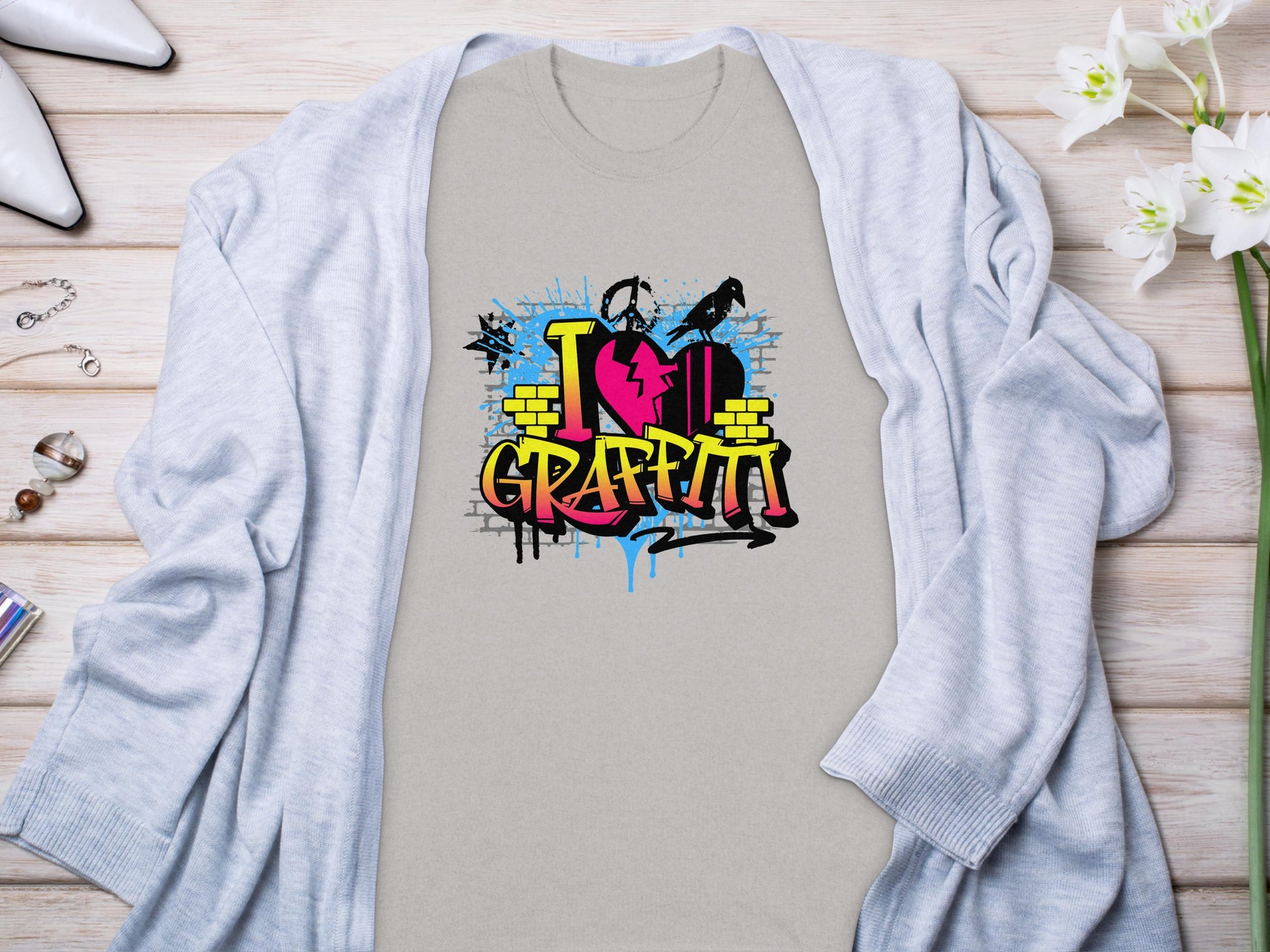 A Garment Graphics light gray Graffiti Love T-Shirt, featuring a vibrant I Love Graffiti street art design, is placed on a wooden surface. It is accompanied by a gray cardigan, white shoes, a bracelet, rings, and white flowers.