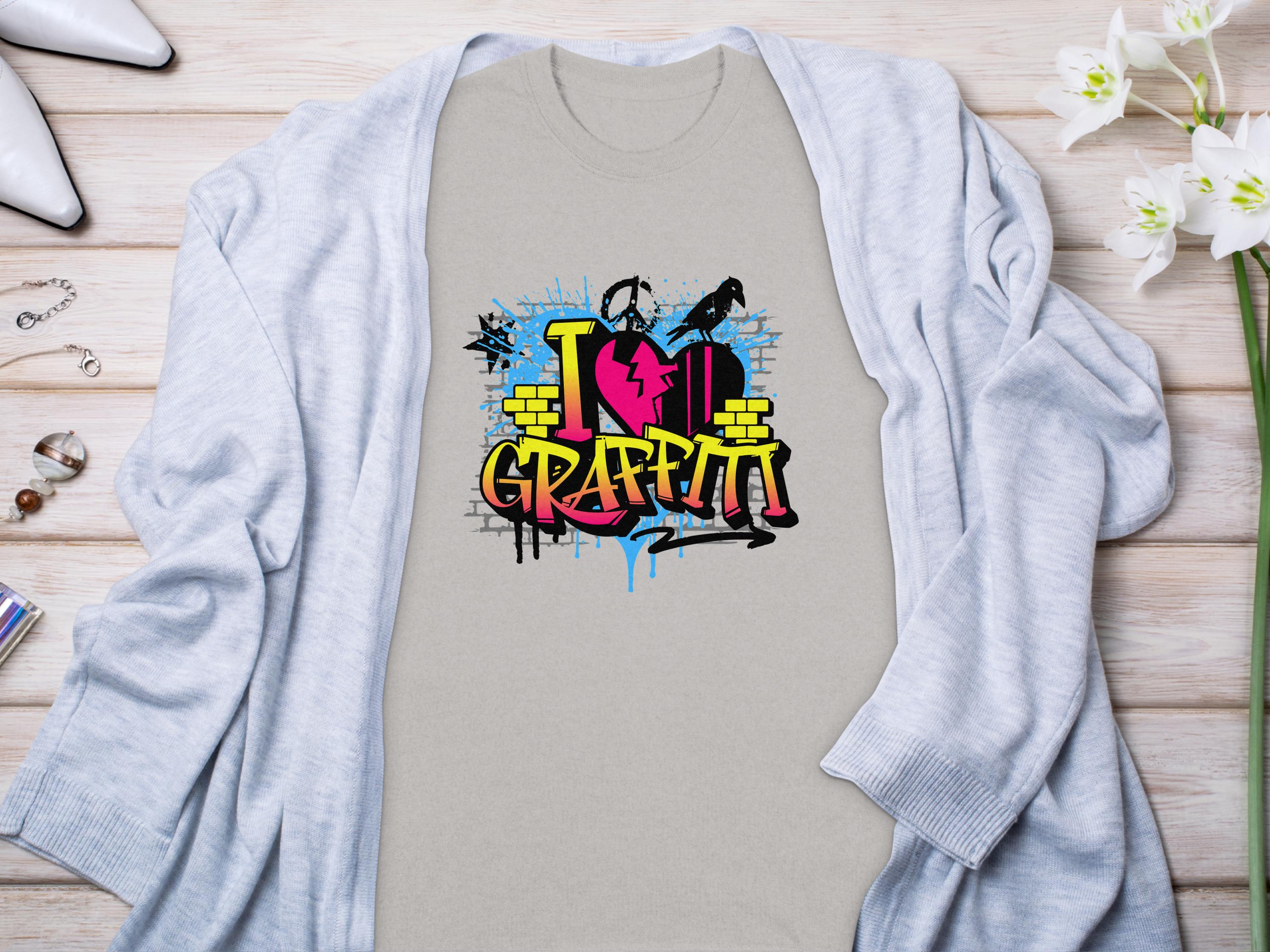 A Garment Graphics light gray Graffiti Love T-Shirt, featuring a vibrant I Love Graffiti street art design, is placed on a wooden surface. It is accompanied by a gray cardigan, white shoes, a bracelet, rings, and white flowers.