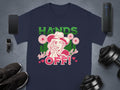 A blue T-shirt by Garment Graphics with a bold cowgirl design holding a gun, surrounded by flowers and hearts. The text reads HANDS OFF! and Wild & Cute. Its in classic fit and is pictured next to a smartphone, smartwatch, speaker, shoes, and dumbbells.