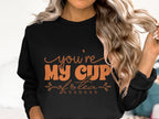 A person in a cozy black Garment Graphics sweatshirt with wavy blonde hair. The unisex sweatshirt features stylized orange text, Youre My Cup of Tea, embellished with leaves and a charming teacup illustration.