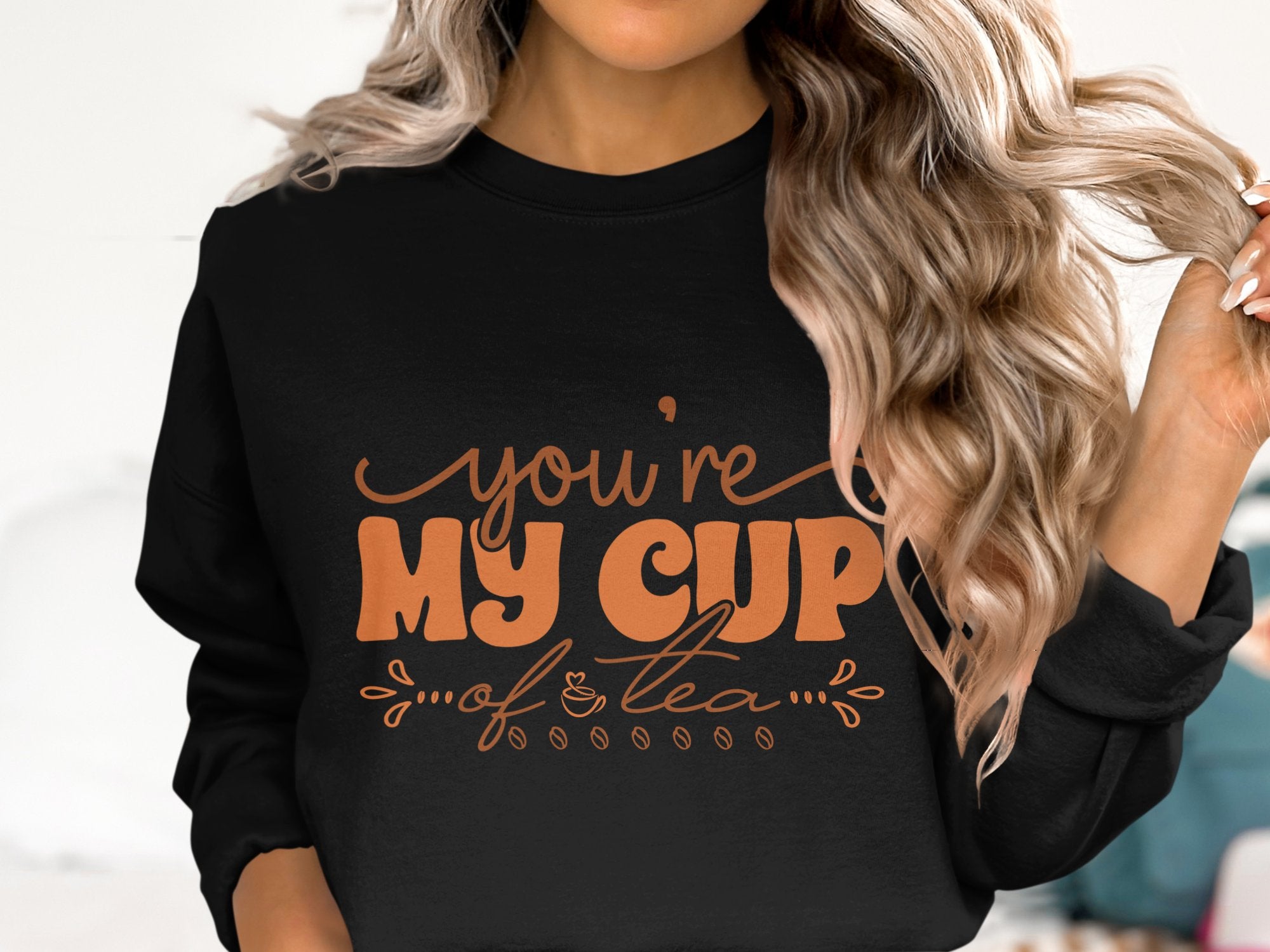 A person in a cozy black Garment Graphics sweatshirt with wavy blonde hair. The unisex sweatshirt features stylized orange text, Youre My Cup of Tea, embellished with leaves and a charming teacup illustration.