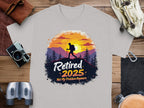 A gray T-shirt by Garment Graphics showcases a hiker with a vibrant sunset and tree frame, featuring Retired 2025: Not My Problem Anymore. Placed on wood alongside essentials like a camera and binoculars, its ideal for those nearing retirement.