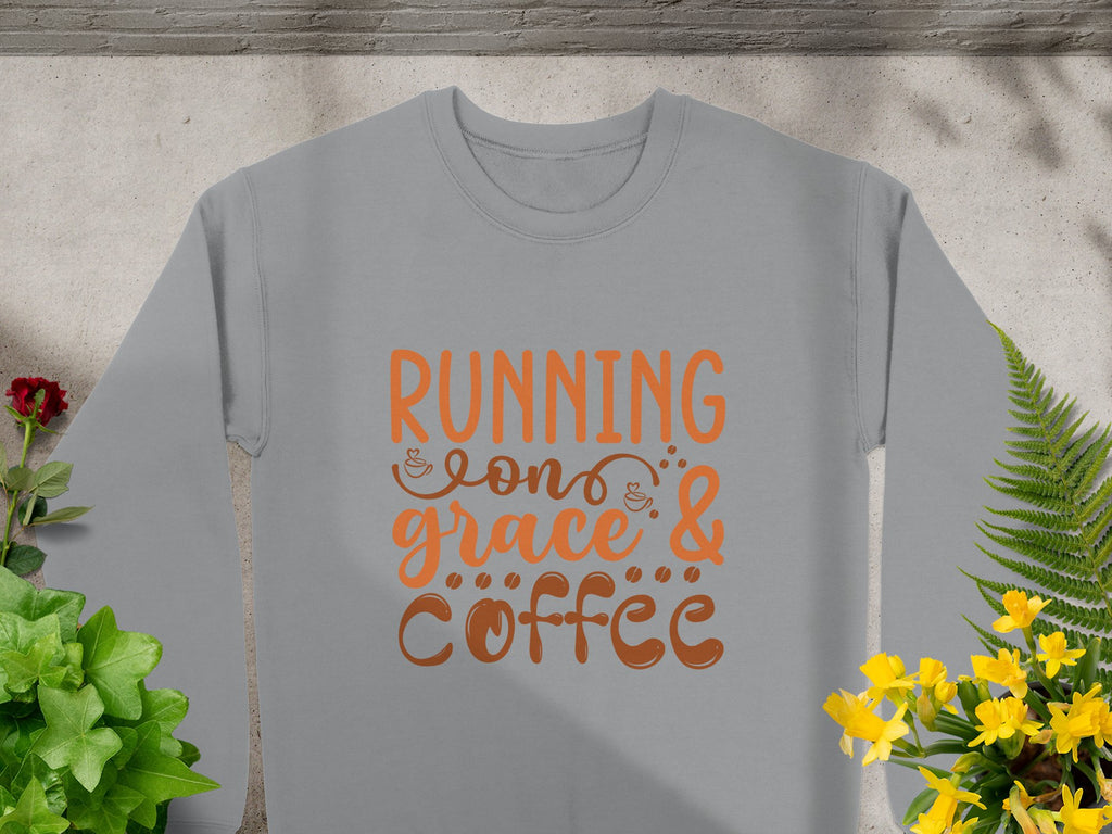 The Garment Graphics sweatshirt showcases Running on Grace & Coffee in vibrant orange. It features a concrete background with shadows, green plants, and colorful flowers. This unisex piece exudes effortless charm.