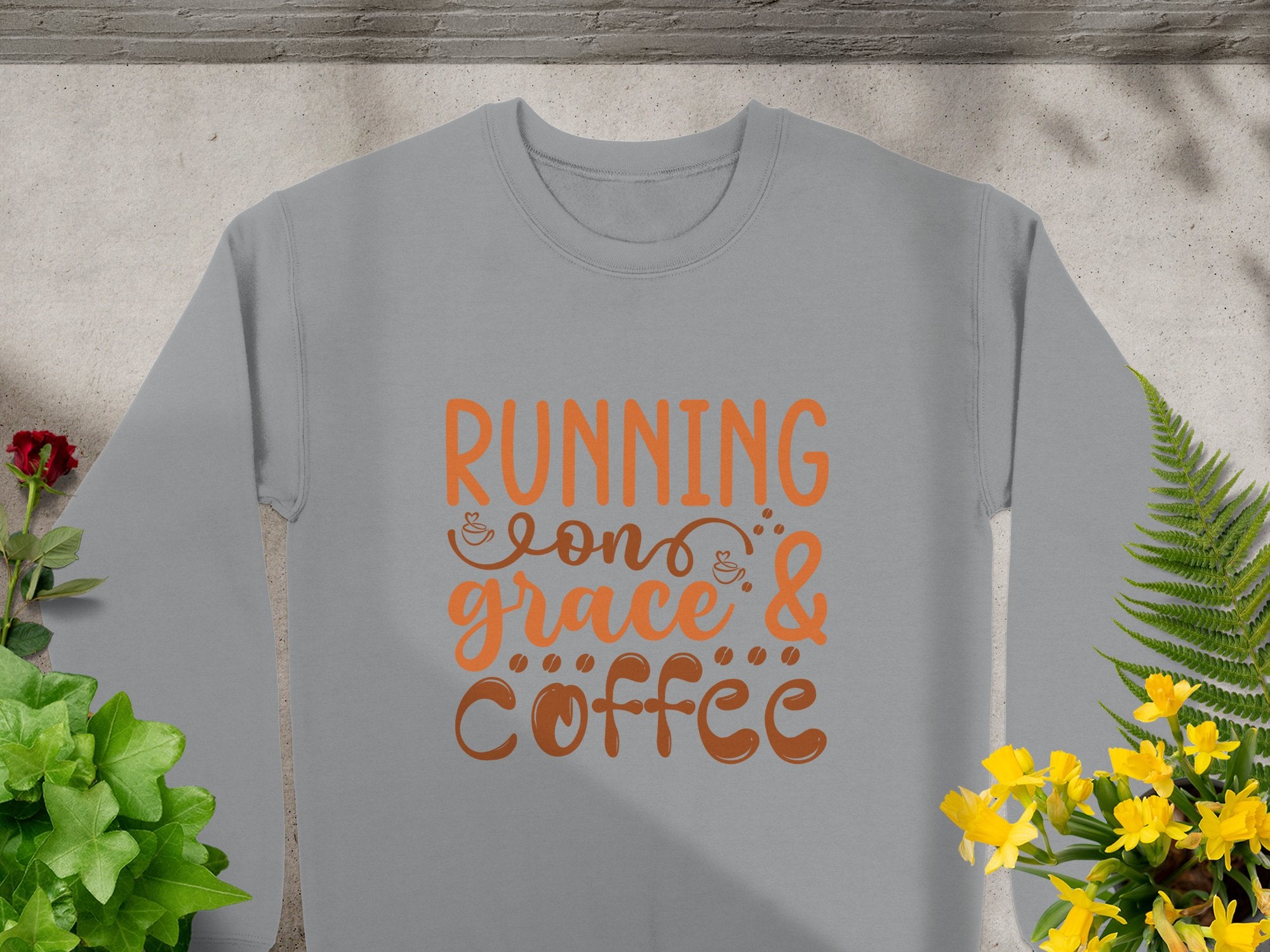 The Garment Graphics sweatshirt showcases Running on Grace & Coffee in vibrant orange. It features a concrete background with shadows, green plants, and colorful flowers. This unisex piece exudes effortless charm.