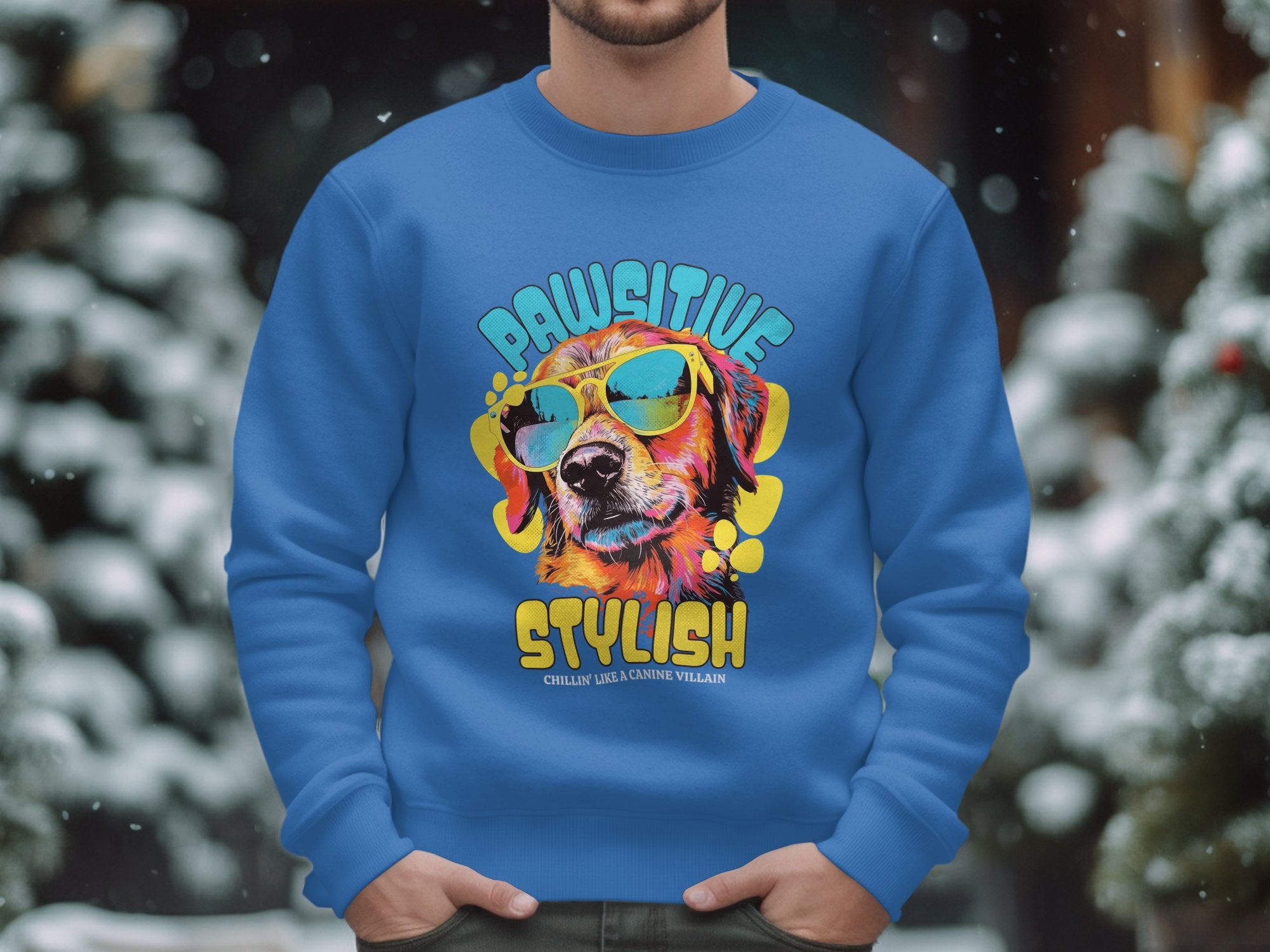 Someone is wearing a blue Garment Graphics sweatshirt featuring a vibrant dog in sunglasses with Pawsitive Stylish and Chillin like a Canine Villain printed below. Snow-covered trees enhance the wintry scene, making it perfect for dog enthusiasts.