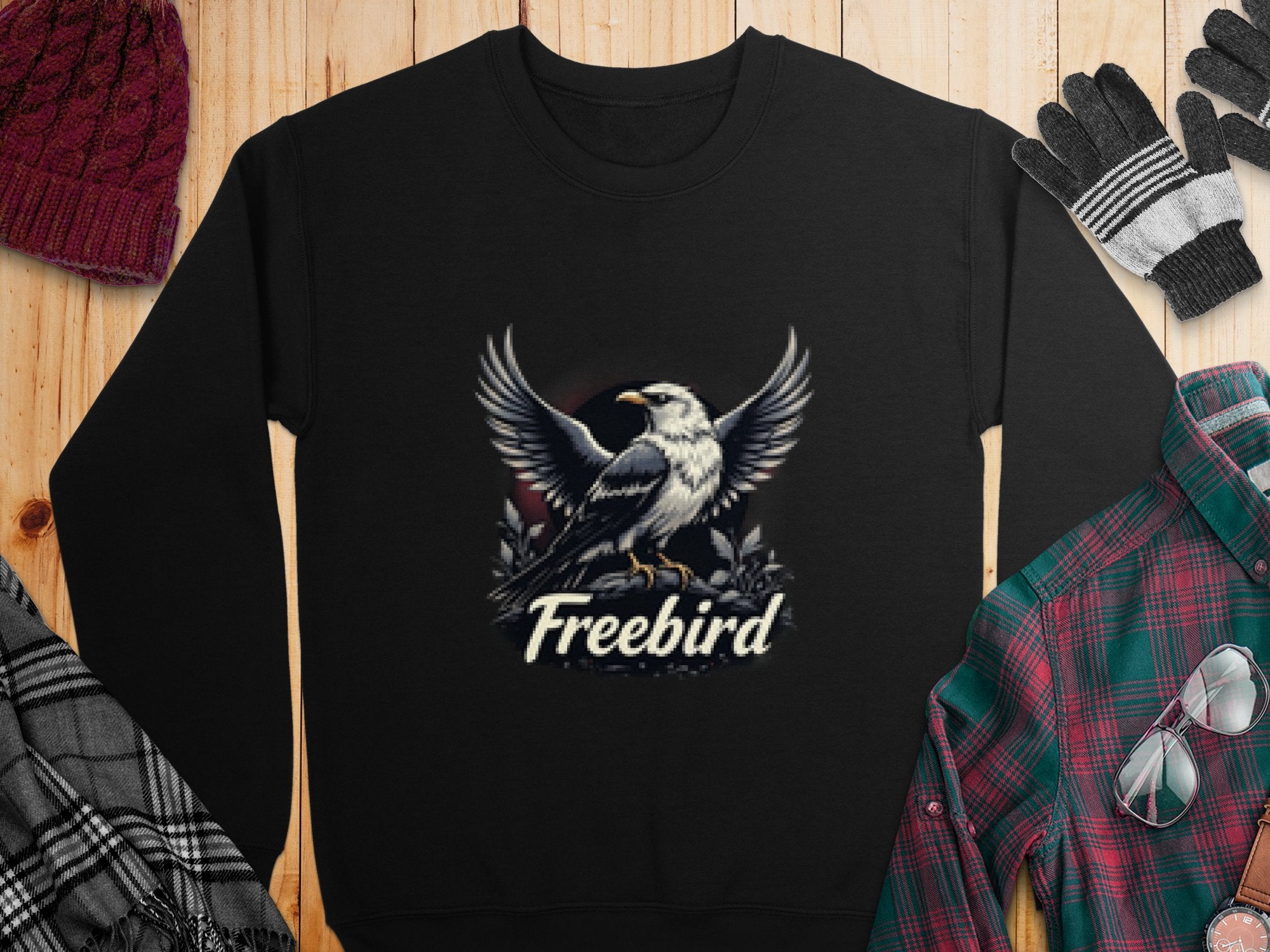 A black Garment Graphics sweatshirt with an eagle and Freebird teams up with a plaid shirt, scarf, knit hat, and gloves on rustic wood, highlighting Freebird Graphic Sweatshirts in unisex sizes.