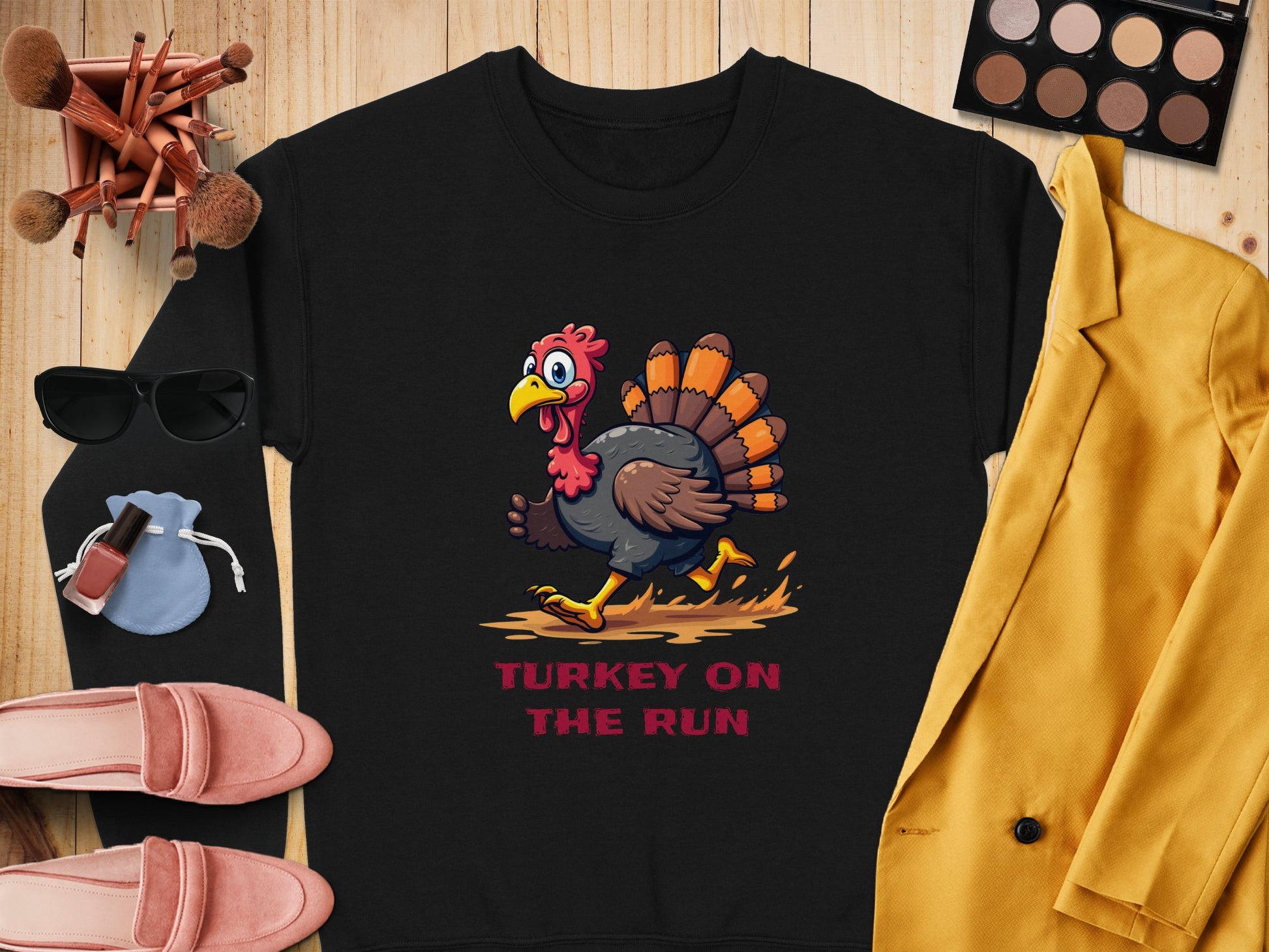 Flat lay featuring Garment Graphics black unisex sweatshirt with a cartoon turkey and TURKEY ON THE RUN, surrounded by a yellow blazer, pink shoes, sunglasses, makeup brushes, an eyeshadow palette, and a light blue pouch with nail polish bottles.