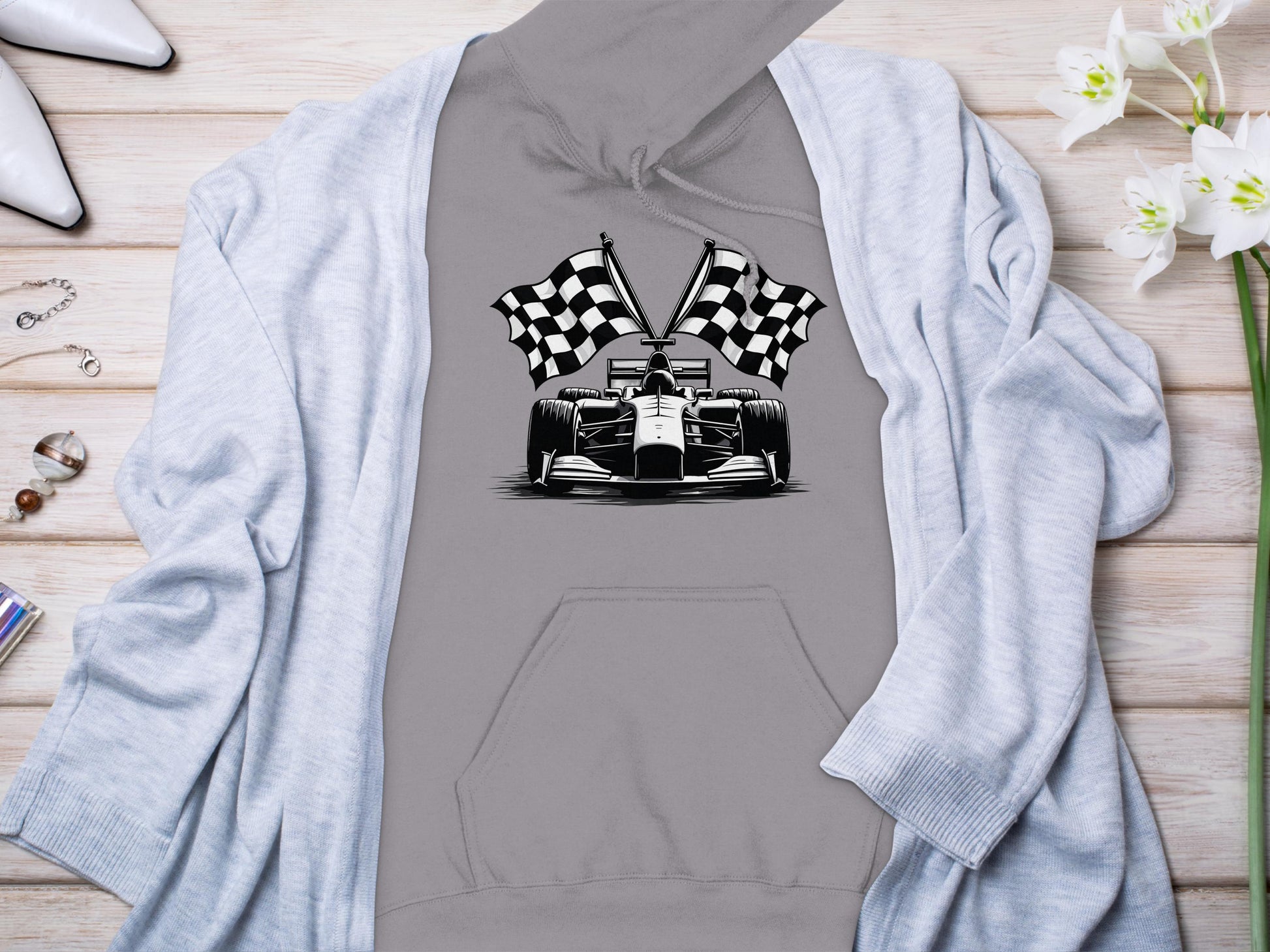 A grey Garment Graphics racing-themed hoodie features a race car graphic with checkered flags on a wooden surface, surrounded by a light blue cardigan, white high-heeled shoes, white flowers, and small jewelry items.